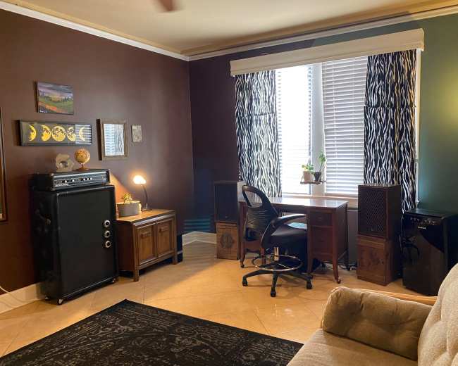 A cozy room with a brown and teal color scheme, featuring a wooden desk, a chair, a record player, and plants near a window with striped curtains.