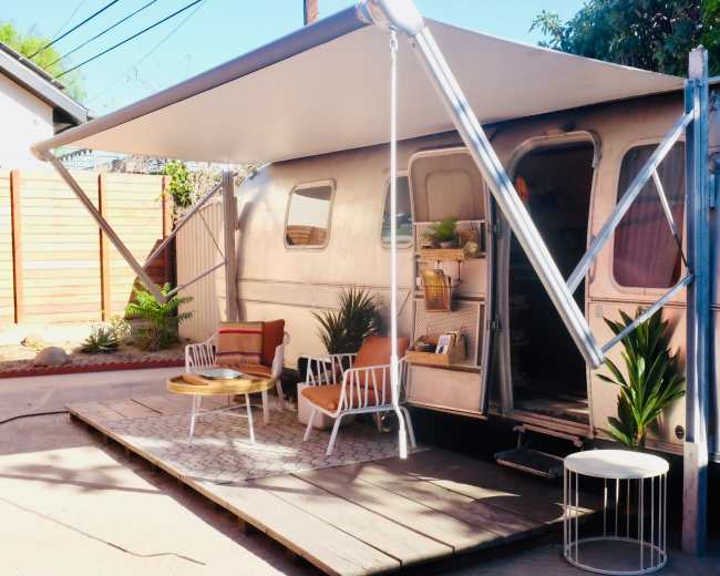 A vintage Airstream trailer with a wooden deck, outdoor seating, and a large awning is positioned near a landscaped area.