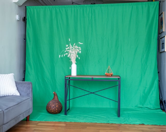 The image features a green backdrop with a table and decorative items in a room that includes a gray sofa and wooden flooring.