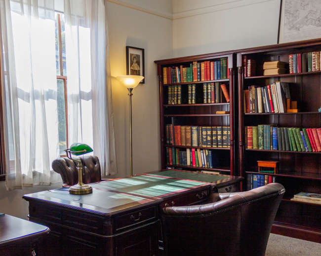 The image depicts a well-furnished office with a desk, a reading lamp, and bookshelves filled with books.
