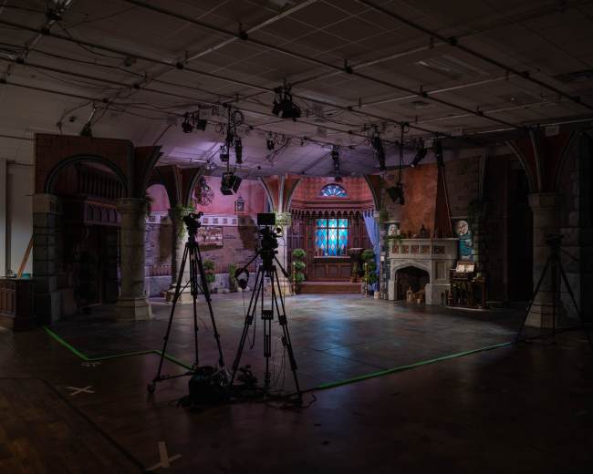 A television studio set features a castle-like interior with cameras positioned around a central stage.