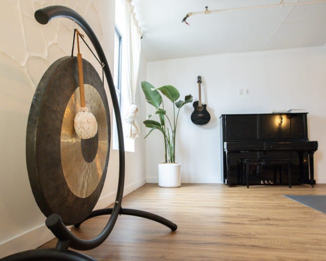 A large gong stands on a black frame in a brightly lit room with a piano and a guitar hanging on the wall.
