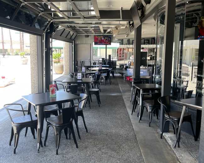 The image shows an empty restaurant interior with black tables and chairs arranged along a walkway, featuring large glass doors that open to the outside.