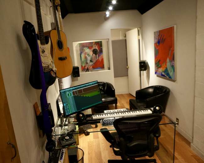 The image shows a small music studio featuring a keyboard, computer, and guitars on the wall, with two black chairs and colorful artwork on display.