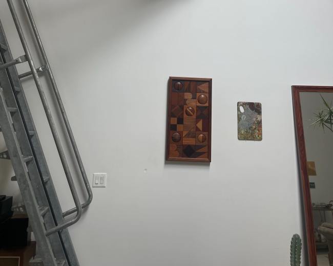 The image shows a minimalist interior with wooden beams, a metal staircase, and two framed artworks on a white wall.