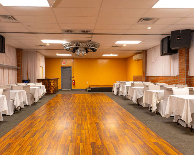 The image shows an empty banquet hall featuring rows of covered tables, wooden flooring, and an orange accent wall.