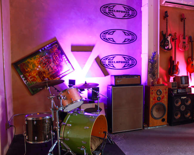 The image shows a music studio corner featuring a green drum set, multiple guitars hanging on the wall, an amplifier, and various audio equipment.