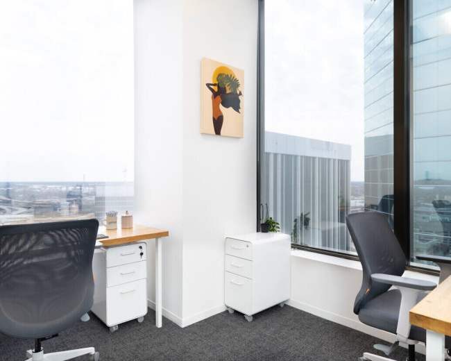 The image shows a modern office space with two desks, a piece of artwork on the wall, and large windows offering a view of the city.