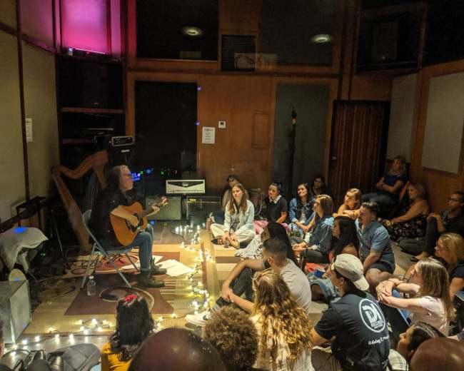A musician plays an acoustic guitar for an attentive audience seated on the floor in a small, dimly lit room.