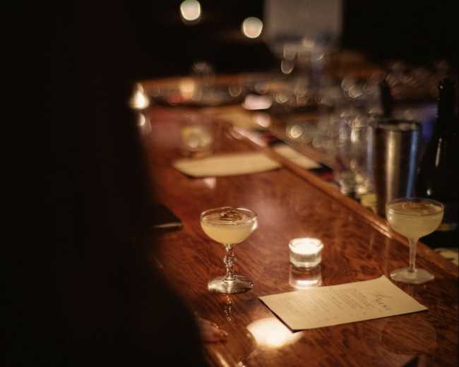 A dimly lit bar is depicted, featuring a long wooden counter with a cocktail glass and a candle placed on top, alongside a menu.
