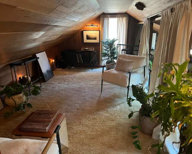 The image shows a cozy attic room with wooden walls, a sloped ceiling, a white armchair, plants, and various decorative elements.