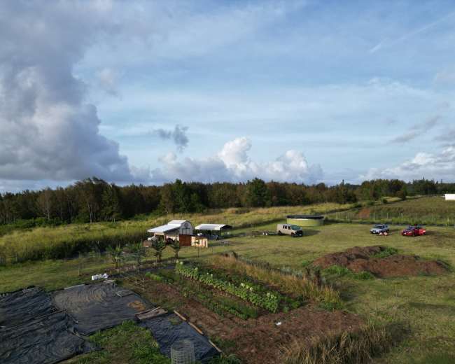 The image shows an aerial view of a rural landscape featuring a small farm with several planted rows, a few vehicles, and structures surrounded by greenery and clouds in the sky.