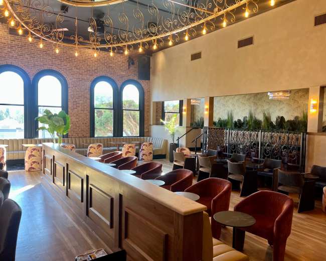 The image shows an upscale lounge with a bar area, plush seating arrangements, large windows, and decorative lighting.