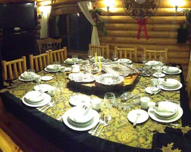 A large, neatly set dining table is arranged in a log cabin, featuring white dishware and a decorative tablecloth, with a view of wooden walls and a television in the background.
