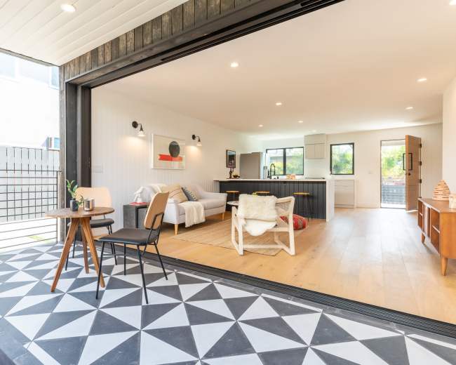 The image shows a modern living space with an open floor plan, featuring a seating area, kitchen, and a patio that opens up to a backyard.