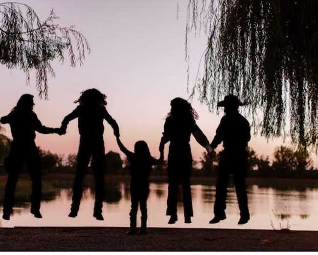 A group of five silhouetted figures, including a child, are holding hands and jumping in front of a lake at sunset.