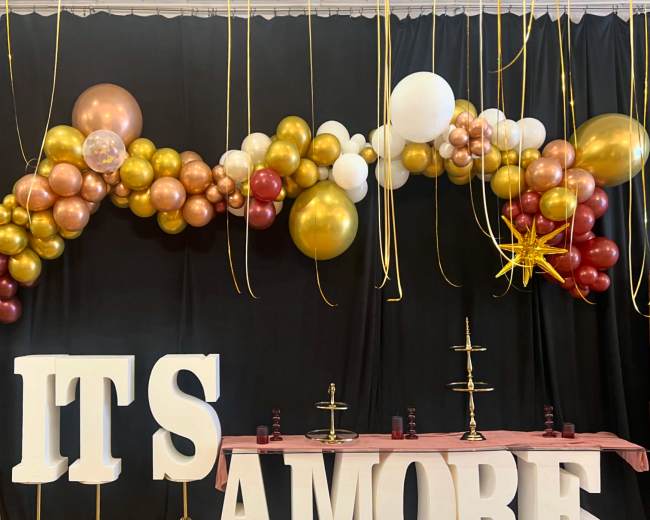 The image features a festive display with a large balloon arch in gold, white, and maroon, and oversized letters spelling "ITS AMORE" on a table with decorative stands beneath a black backdrop.