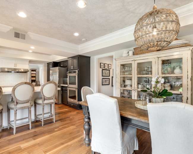 The image shows a kitchen with a wooden dining table and chairs, an island bar with stools, and appliances integrated into the cabinetry.
