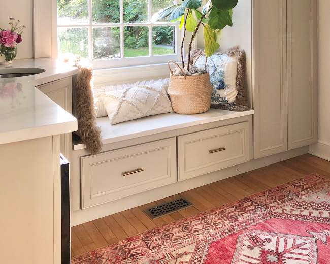 A window seat with decorative cushions and a potted plant is situated beside a large window, illuminated by natural light.