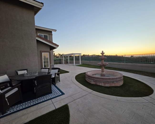 The image shows a landscaped outdoor patio with a fountain and seating area, surrounded by greenery under a clear sky at sunset.