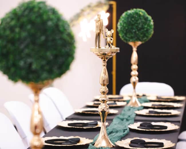 A long dining table is set with gold-rimmed plates, black napkins, and topped with gold candlesticks and small green topiaries.
