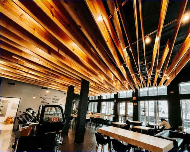 The interior of a modern café features a ceiling with wooden beams and pendant lights, large windows, and wooden tables.