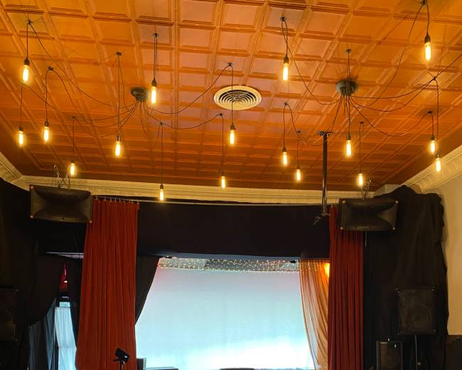 The image depicts a performance stage in a venue, featuring a red curtain backdrop, wooden flooring, and suspended light bulbs overhead.