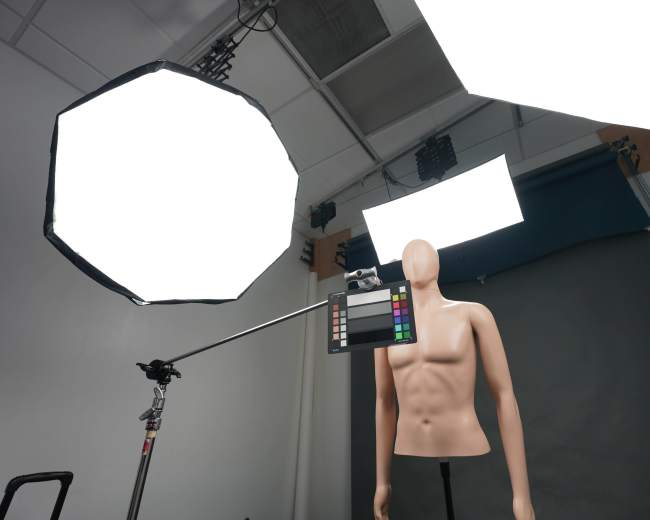 A photography studio setup features a mannequin in front of a gray backdrop, with softbox lights positioned overhead and a color reference chart attached to a camera on a tripod.