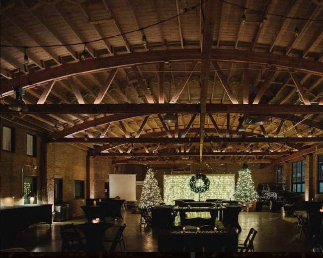 A spacious event venue features wooden beams, festive decorations, and tables arranged for a gathering, with Christmas trees and lights adorning the space.