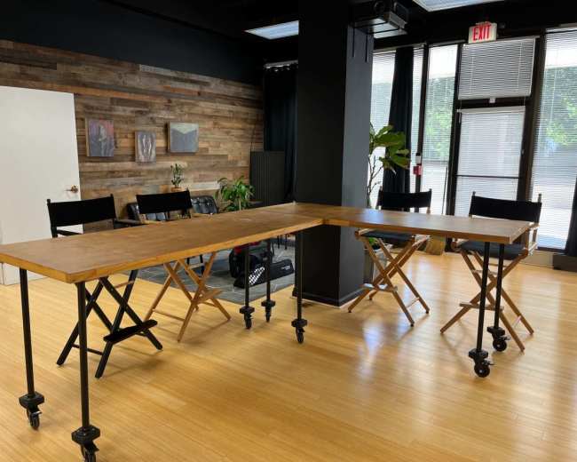 A long wooden table with metal legs is positioned at an angle in a room with hardwood flooring, accompanied by several black director's chairs.