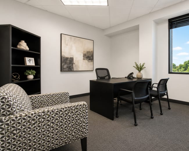 The image shows a modern office space furnished with a black desk and three chairs, a patterned armchair, a black bookshelf, and a large window overlooking greenery.