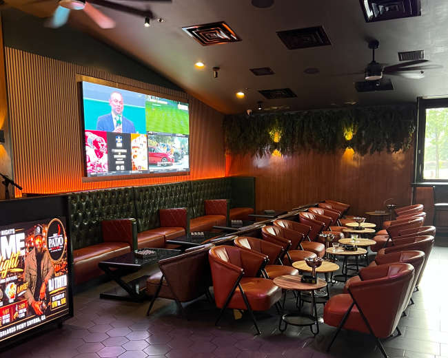 The image shows a spacious lounge area with red chairs and low wooden tables, featuring a large screen displaying a sports event on the wall.
