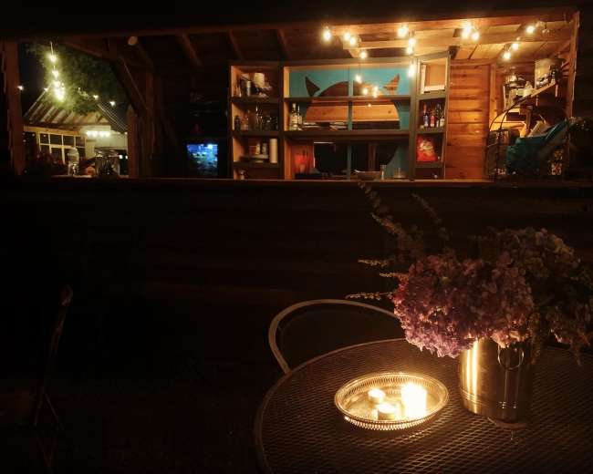 A cozy outdoor setting with a table featuring a flower vase and candles, illuminated by string lights hung above a rustic bar area in the background.