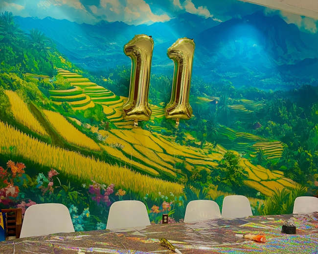 A colorful party table is set against a vibrant mural of rice terraces and mountains, with gold number balloons reading "11" above.