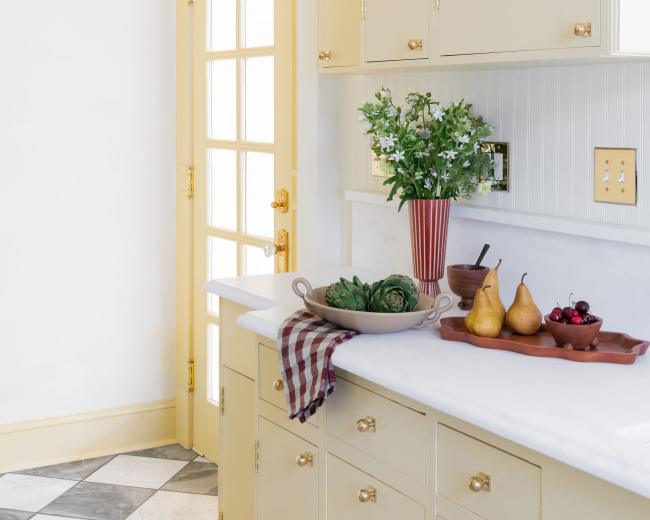 The image shows a light yellow kitchen with a marble countertop, a bouquet of greenery in a vase, and a wooden tray featuring fruits, with a door leading to the outside.