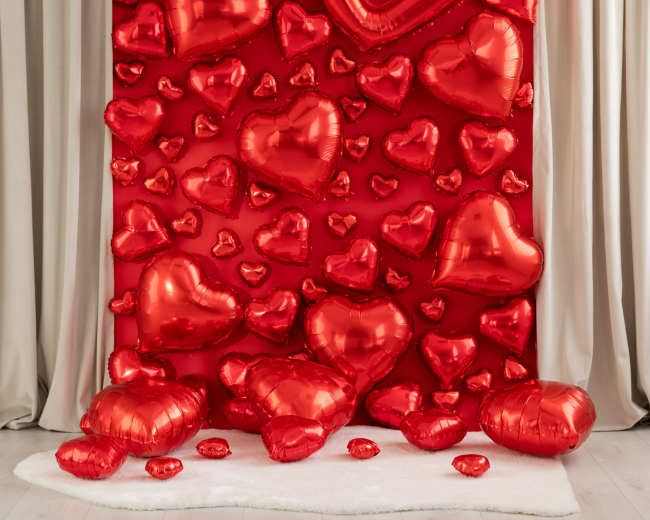 The image features a backdrop decorated with red heart-shaped balloons arranged on a red wall, with additional inflated hearts scattered on the floor.