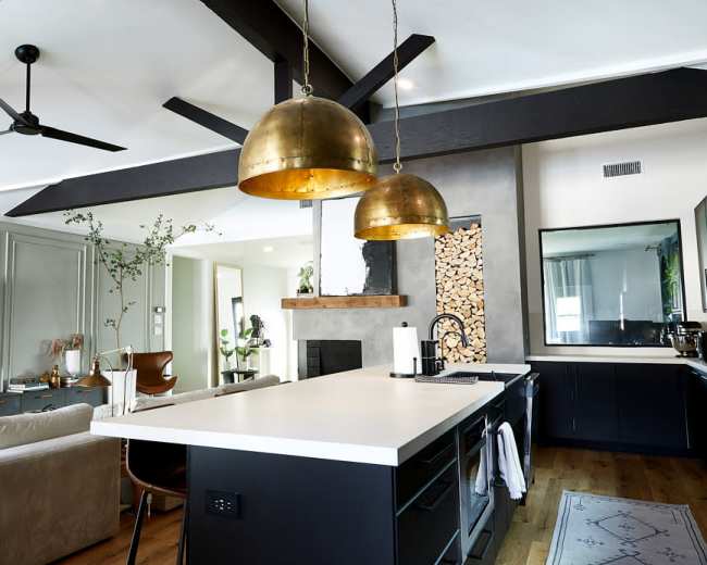 The image shows a modern kitchen and living room area featuring a large island with a white countertop, two brass pendant lights, and a wooden beam ceiling.