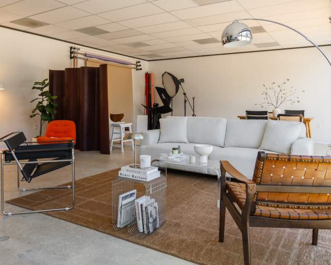 The image shows a modern, spacious interior with a light-colored sofa, an orange chair, a wooden dining table, and various photography equipment in the background.