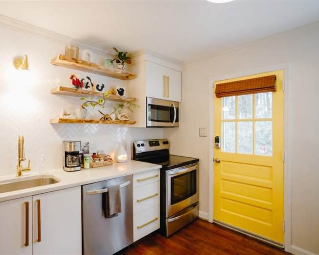 A small kitchen features open wooden shelves, stainless steel appliances, and a bright yellow door.
