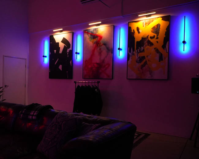 The image shows a dimly lit room featuring three large abstract paintings on the wall, illuminated by blue LED lights, with a sectional leather couch in the foreground.