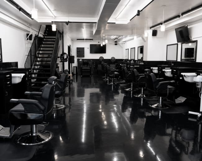 The image shows a modern, spacious hair salon with black styling chairs, mirrors, and sinks arranged neatly along the walls and rows of lights above.