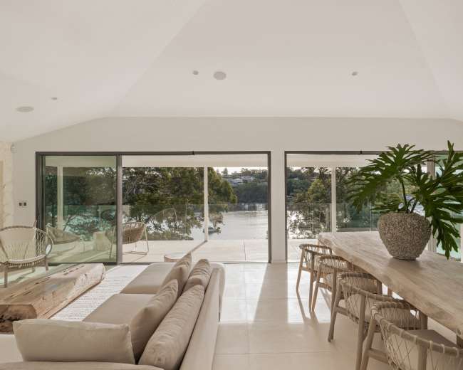 The image shows a modern living space with large glass doors that open to a waterfront view, featuring a beige sofa, a wooden dining table, and a large potted plant.