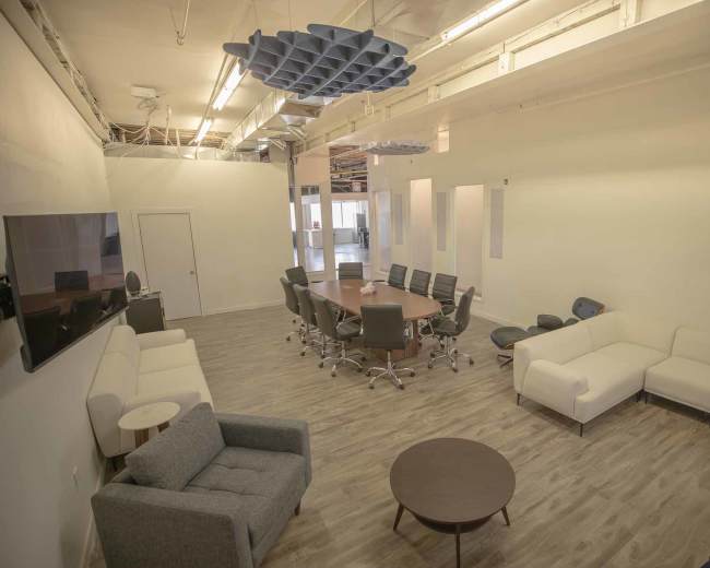 The image shows a modern office space with a large conference table surrounded by black chairs, complemented by sectional sofas and a coffee table in a neutral-toned room.