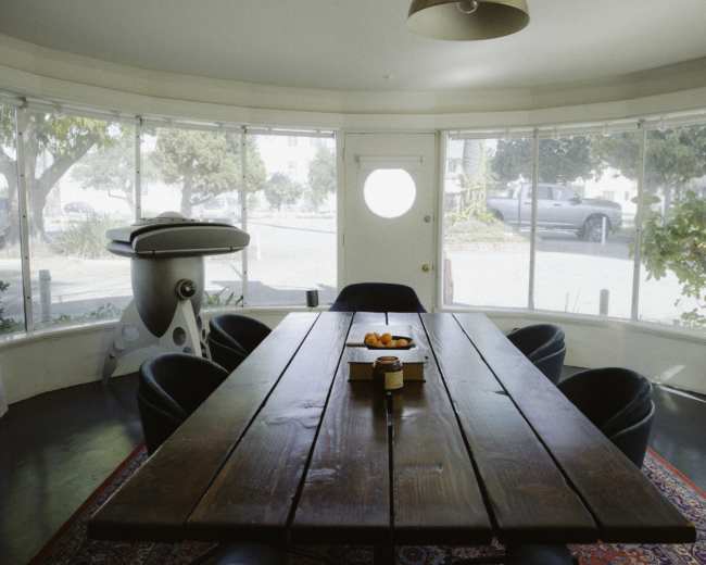 A large wooden dining table surrounded by black chairs is positioned in a bright, circular room with large windows and a door that opens to the exterior.