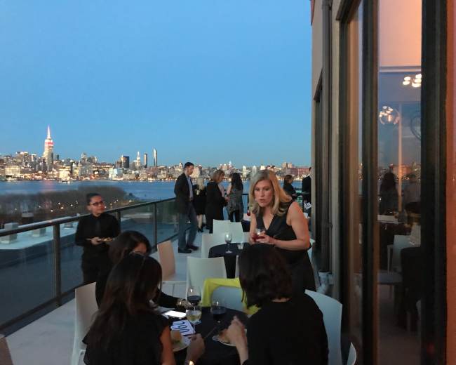 A group of people socializes on a rooftop terrace overlooking the Manhattan skyline at dusk.