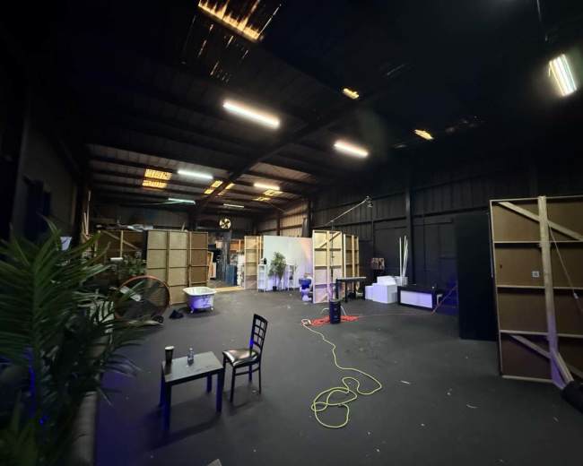 The image shows an industrial space with a dark floor and walls, featuring wooden frames, a few plants, and scattered furniture in a partially lit environment.