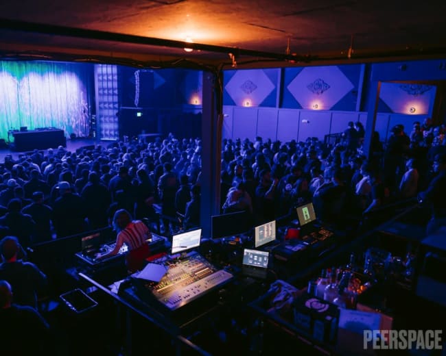A large crowd is gathered in a dimly lit venue facing a stage with a shimmering backdrop and audio equipment set up in the foreground.