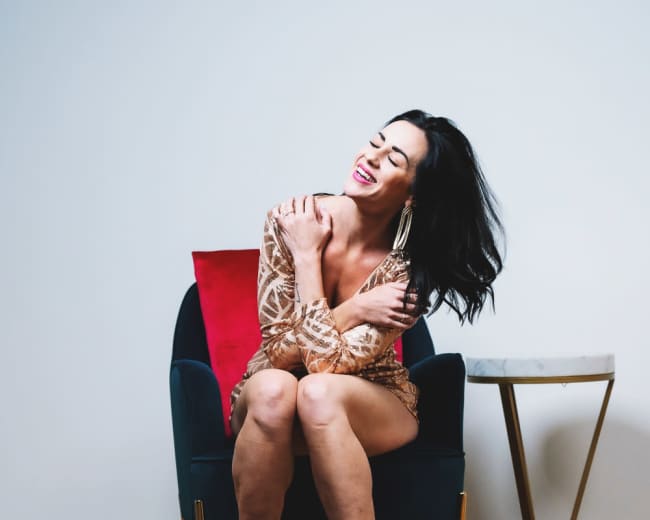 A woman sitting in a chair smiles while hugging her arms, with a colorful rug and a table nearby.