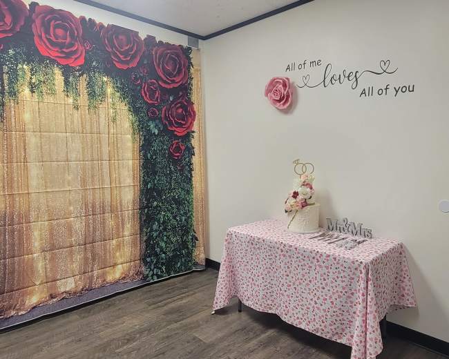 A decorated room featuring a large floral wall backdrop, a table covered with a patterned cloth, and a wall inscription that reads "All of me loves All of you."