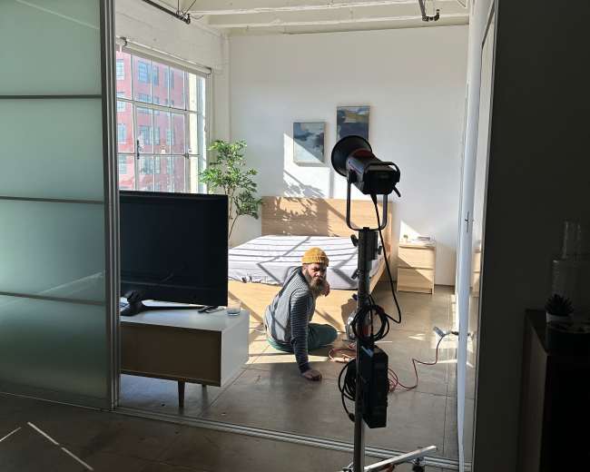 A person sits on the floor of a well-lit room with a bed and a television, while a lighting setup is positioned nearby.